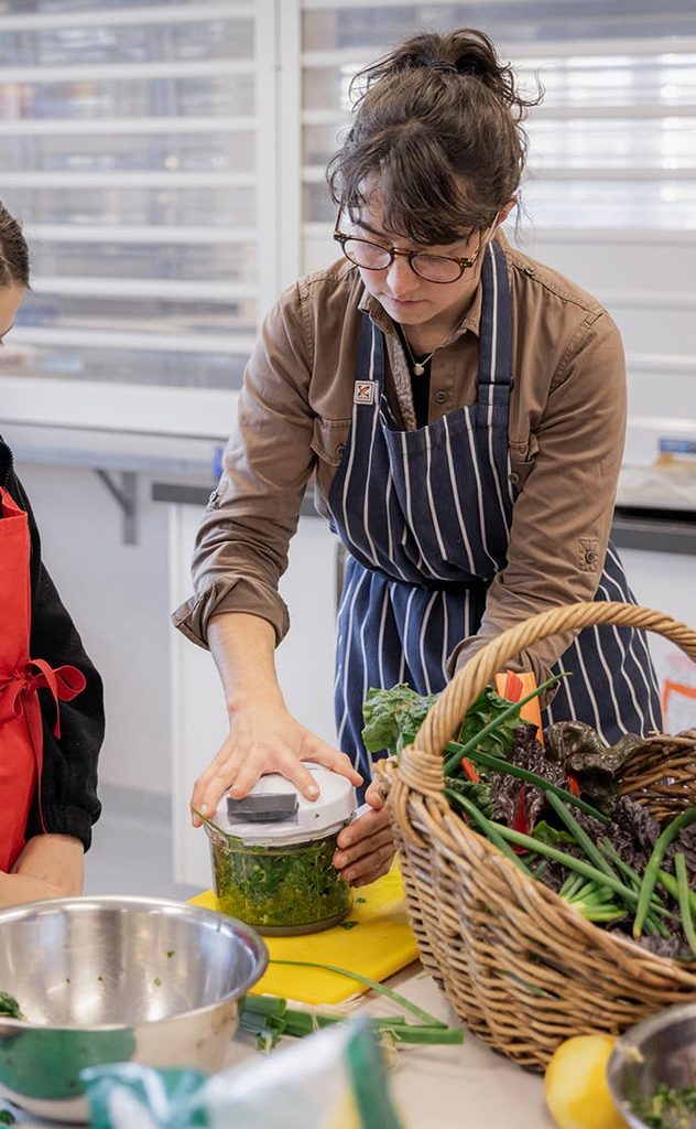 A teacher is standing next to a student while they blend herbs.